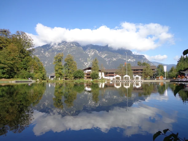Riesserseehotel mit Kramergebirge Hotel am See mit Bergkulisse, spiegelndem Wasser und umliegenden Bäumen unter blauem Himmel.