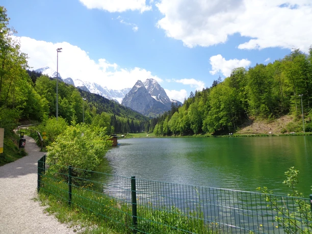 Uferweg mit Blick auf den kleinen und großen Waxenstein Uferweg am klaren See mit bewaldeten Hängen und schneebedeckten Gipfeln im Hintergrund.