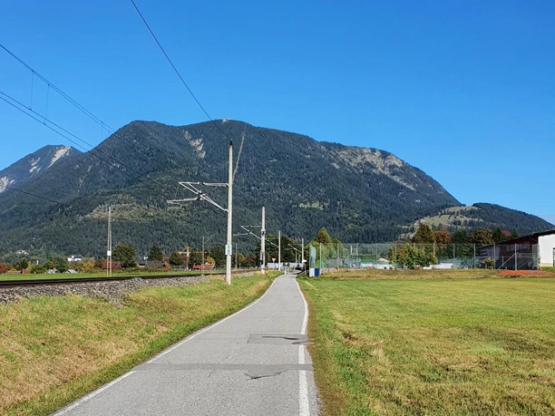 Weg zurück zum Ausgangspunkt Gerader Weg zwischen Wiese und Gleisen vor bewaldetem Berg unter klarem blauem Himmel.