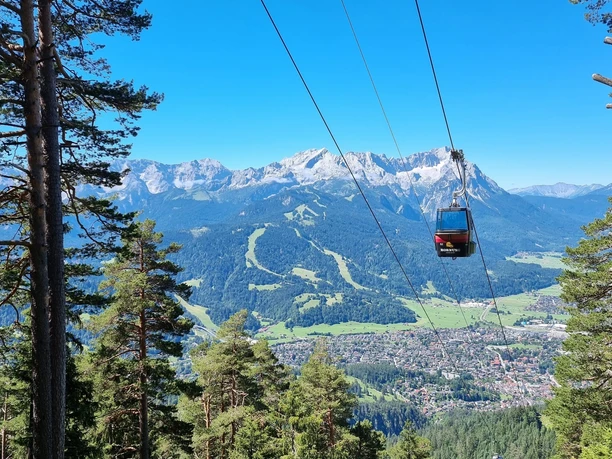 Wankbahn Seilbahn über bewaldetem Hang mit weitem Blick auf Tal und Bergmassiv unter klarem Himmel.
