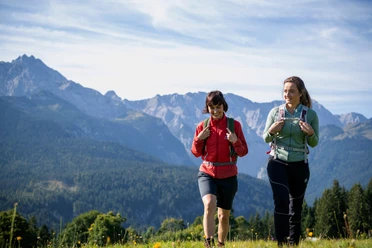 Wandern mit Ausblick Zwei Menschen wandern vor hohen Bergen. Du siehst grünes Gras und klare Luft.