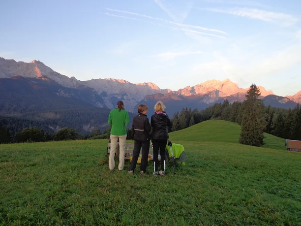 Sonnenaufgangstour Eckbauer Drei Personen blicken von einer Wiese auf sonnenbeschienene Berge im frühen Morgenlicht