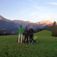 Am Eckbauer Drei Personen stehen auf einer Wiese und blicken auf Berge im warmen Abendlicht