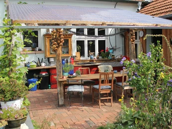Overdekte buitenkeuken met rustieke houten tafel, stoelen en planten in de tuin.
