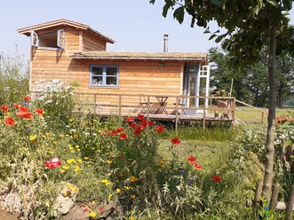 Tiny Haus Storchennest Holzhaus auf einer Veranda mit bunten Blumen, umgeben von grüner Landschaft und klarem Himmel.