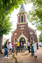 Gruppe von Menschen steht vor einer Backsteinkirche mit hohem Turm, umgeben von Bäumen im Clara Ernst Platz.