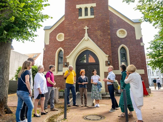 Gruppe von Menschen steht vor einer Backsteinkirche mit hohem Turm, umgeben von Bäumen im Clara Ernst Platz.