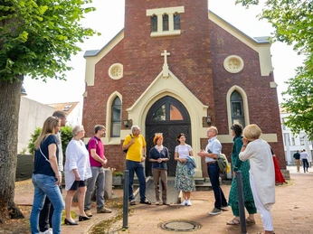 Clara Ernst Platz Gruppe von Menschen steht vor einer Backsteinkirche mit hohem Turm, umgeben von Bäumen im Clara Ernst Platz.