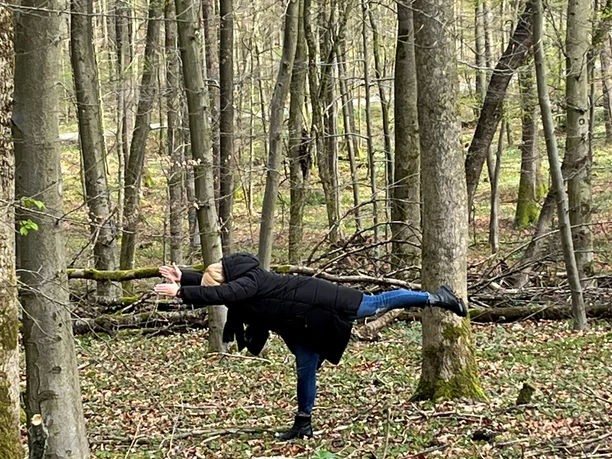 Eine Frau in schwarzem Mantel macht Yoga-Pose im grün-braunen Herbstwald, umgeben von Bäumen.