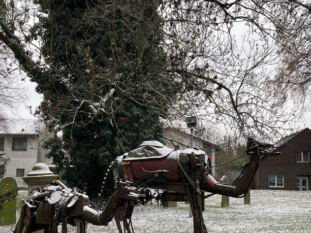 Ein Kamel und Reiter aus Metall stehen auf einer verschneiten Wiese, umgeben von Bäumen.