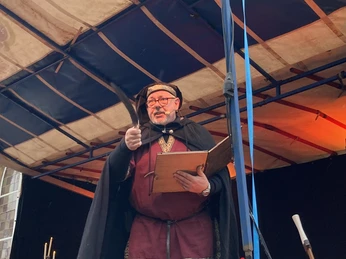 Schauspieler in mittelalterlicher Kleidung liest auf einer festlichen Bühne aus einem Buch vor.