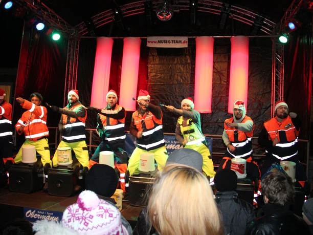 Eine Gruppe von Männern in farbenfrohen Outfits und Weihnachtsmützen tanzt energisch auf einer beleuchteten Bühne.