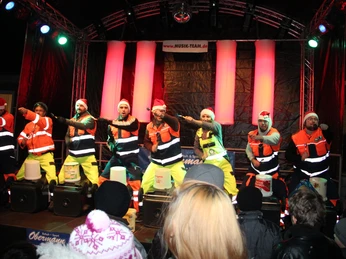 Dezemberträume Rahden Eine Gruppe von Männern in farbenfrohen Outfits und Weihnachtsmützen tanzt energisch auf einer beleuchteten Bühne.