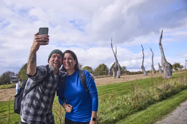 Försterteiche-StaffPark-LTMGmbH-HirschmeierMediaGmbH&CoKG_076.jpg Ein lächelndes Paar macht ein Selfie vor einer Skulpturenlandschaft mit markanten Bäumen.