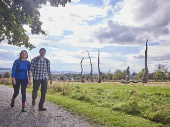 Zwei Personen spazieren auf einem Schotterweg durch eine weitläufige Landschaft mit Skulpturen.