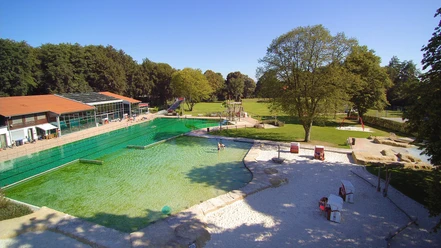 Parkbad.jpg Freizeitpark mit grünem Schwimmbecken, Sitzgelegenheiten und großer Wiese unter klarem Himmel.
