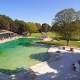 Parkbad.jpg Freizeitpark mit grünem Schwimmbecken, Sitzgelegenheiten und großer Wiese unter klarem Himmel.