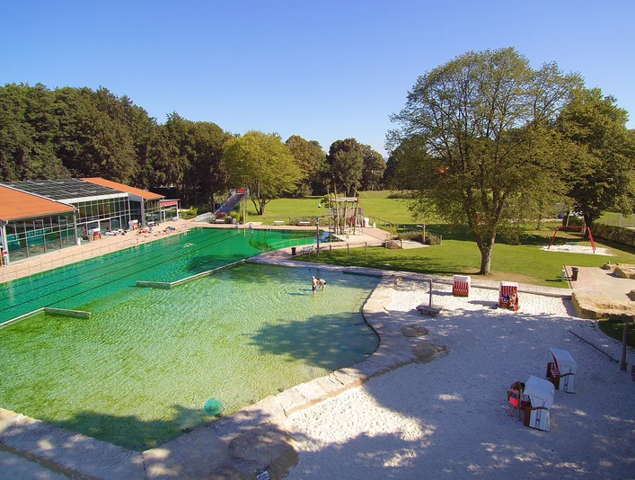 Parkbad.jpg Freizeitpark mit grünem Schwimmbecken, Sitzgelegenheiten und großer Wiese unter klarem Himmel.