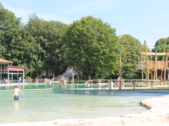 Barrierefreies Freibad mit natürlichem Schwimmbecken, Holzspielbereich und umgeben von grünen Bäumen.
