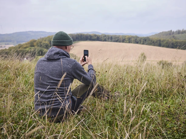Wilddiebsroute-LTMGmbH-HirschmeierMediaGmbH&CoKG_082.jpg Mann in grauem Hoodie fotografiert mit Smartphone eine weitläufige, hügelige Landschaft.