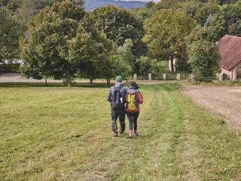 Zwei Personen wandern auf einem grasbewachsenen Weg, umgeben von Bäumen und einem Gebäude.
