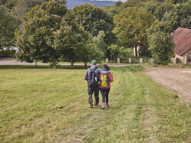 Wilddiebsroute-LTMGmbH-HirschmeierMediaGmbH&CoKG_084.jpg Zwei Personen wandern auf einem grasbewachsenen Weg, umgeben von Bäumen und einem Gebäude.