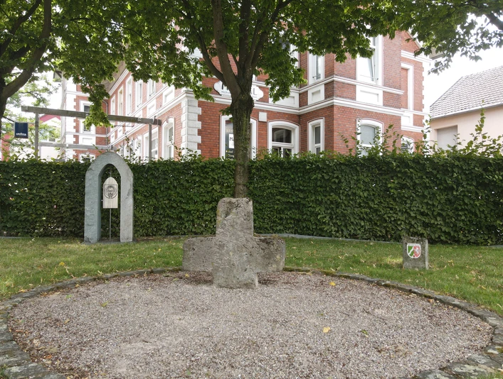DJI_0396.JPG Ein steinernes Kreuzdenkmal unter Bäumen, nahe eines historischen Backsteingebäudes und eines Wappens.