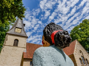 Bockhorst Dorfkirche.jpeg Steinfigur einer Frau mit rotem Haarband vor einer Kirche mit Uhrturm, Baum und teils bewölktem Himmel.