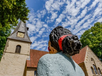 Bockhorst Dorfkirche.jpeg Steinfigur einer Frau mit rotem Haarband vor einer Kirche mit Uhrturm, Baum und teils bewölktem Himmel.