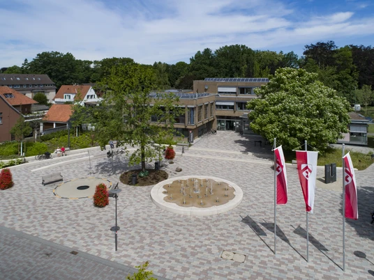 Luftbild Rathaus tiefer 032 © Markus Stein.jpg Moderner Platz mit Springbrunnen, drei rot-weißen Fahnen, umgeben von Bäumen und Backsteinhäusern.