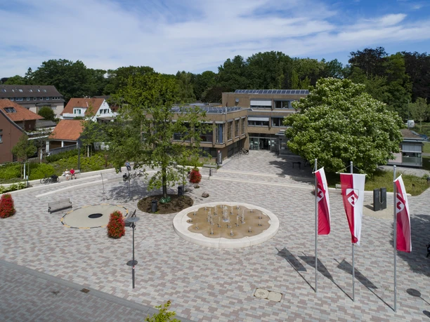Luftbild Rathaus tiefer 032 © Markus Stein.jpg Moderner Platz mit Springbrunnen, drei rot-weißen Fahnen, umgeben von Bäumen und Backsteinhäusern.
