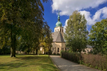Park bij de Lutherse kerk Peter-Paul Een kerk met een groene toren staat tussen bomen onder een blauwe hemel; een geplaveid pad leidt ernaartoe.