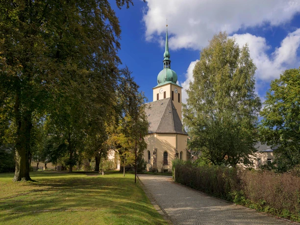 Parkanlage mit Ev. Luth. Kirche Peter-Paul Eine Kirche mit grünem Turm steht zwischen Bäumen unter blauem Himmel; ein gepflasterter Weg führt dorthin.
