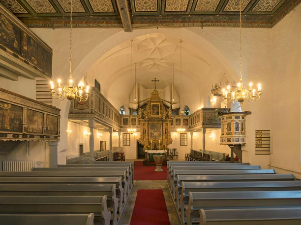 Innenansicht-Blick zum Altar Innenansicht einer Kirche mit Holzbänken, rotem Teppich, verziertem Altar und Kronleuchtern; warme, einladende Atmosphäre.