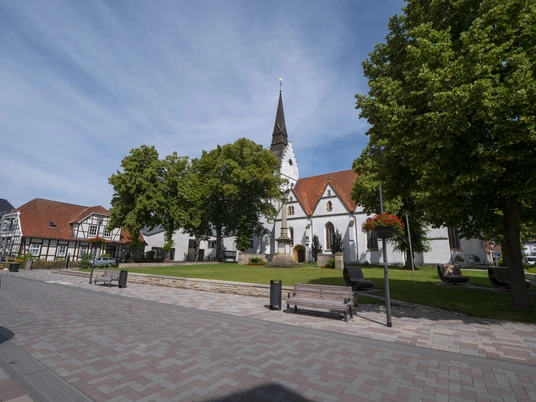 Kirche St. Petri 088 @ Markus Stein.jpg Historische Kirche mit spitzem Turm umgeben von grünen Bäumen und Sitzbänken in einer Fußgängerzone.