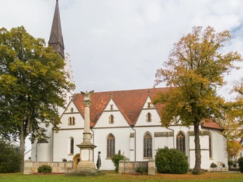 Eine weiße Kirche mit spitzem Turm, umgeben von Bäumen, im Herbstlaub, vor bewölktem Himmel.
