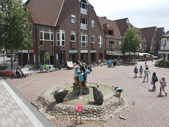 Kinder spielen auf einem Brunnen mit Wildschweinskulpturen im Zentrum einer gepflasterten Platzes.