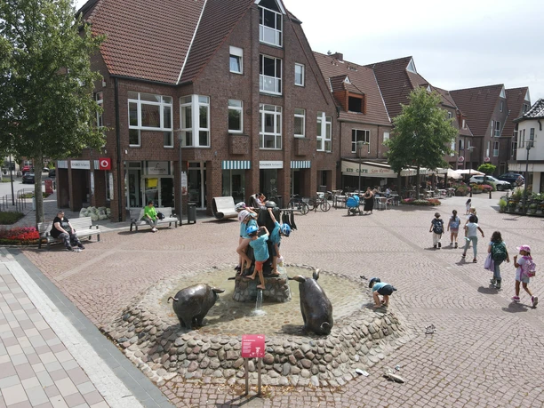 Kinder spielen auf einem Brunnen mit Wildschweinskulpturen im Zentrum einer gepflasterten Platzes.