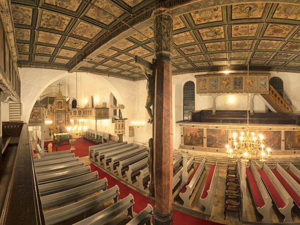 Interior view from the gallery Interior view of a church with painted wooden ceiling, red carpets and gray wooden benches; a chandelier hangs in the center.