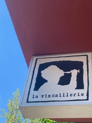 La Vincaillerie Ein quadratisches Schild zeigt das stilisierte Profil einer Person, die aus einem Weinglas trinkt, mit der Aufschrift "la vincaillerie" darunter, auf rotem Gebäudehintergrund vor blauem Himmel.