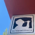 La Vincaillerie Ein quadratisches Schild zeigt das stilisierte Profil einer Person, die aus einem Weinglas trinkt, mit der Aufschrift "la vincaillerie" darunter, auf rotem Gebäudehintergrund vor blauem Himmel.A square sign shows the stylized profile of a person drinking from a wine glass, with the inscription "la vincaillerie" underneath, on a red building background against a blue sky.