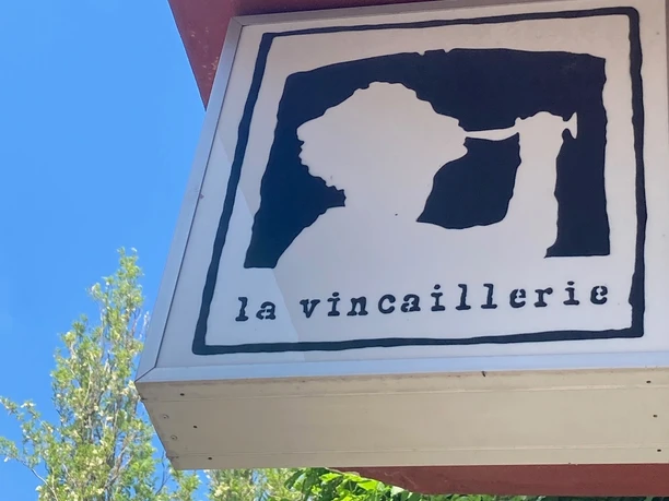 La Vincaillerie A square sign shows the stylized profile of a person drinking from a wine glass, with the inscription "la vincaillerie" underneath, on a red building background against a blue sky.