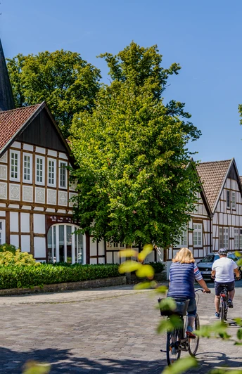 Fachwerkhäuser in sonniger Dorfstraße, umgeben von grünen Bäumen, zwei Radfahrer fahren gemütlich vorbei.