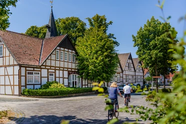 Bockhorst Radfahrer.jpeg Fachwerkhäuser in sonniger Dorfstraße, umgeben von grünen Bäumen, zwei Radfahrer fahren gemütlich vorbei.