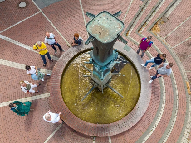 Brunnen in Kreisform aus Ziegelsteinen, zentrale Bronzeskulptur, umgeben von flanierenden Menschen.