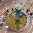 Zieglerbrunnen Brunnen in Kreisform aus Ziegelsteinen, zentrale Bronzeskulptur, umgeben von flanierenden Menschen.
