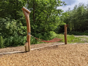 Rote Netz-Hängematte hängt zwischen zwei Holzpfosten auf einem Boden aus Mulch mit grüner Waldkulisse.