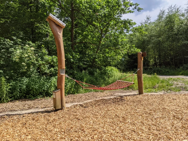 Hängematte Rote Netz-Hängematte hängt zwischen zwei Holzpfosten auf einem Boden aus Mulch mit grüner Waldkulisse.