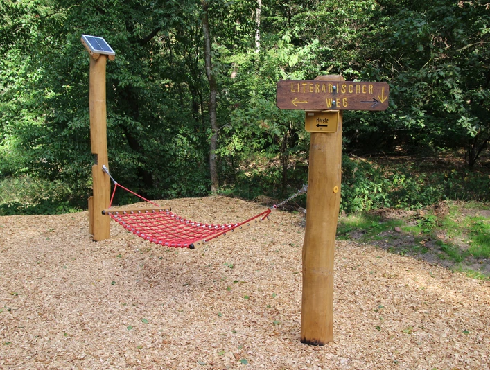 Rote Hängematte aus robustem Material im Wald, flankiert von Holzstangen und Wegweiser "Literarischer Weg".