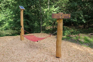Hängematte Rote Hängematte aus robustem Material im Wald, flankiert von Holzstangen und Wegweiser "Literarischer Weg".